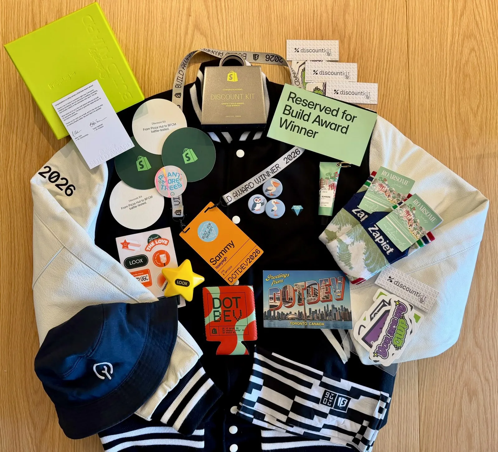 The 2026 Build Award haul laid out on a table: a black-and-cream varsity jacket with 2026 on the sleeve, a gold box reading “Congratulations Discount Kit — Shopify Build Award 2026 Winner”, Build Award lanyards, a “Reserved for Build Award Winner” card, a DotDev Toronto postcard, and a pile of discountkit stickers